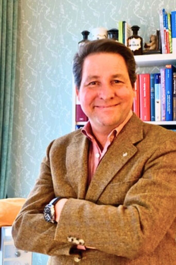 Heilpraktiker Marco Beitelberger steht freundlich lächelnd in seiner Naturpraxis Wien 1140 vor einem Bücherregal.
