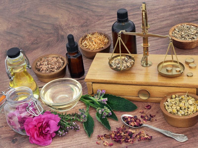 Apothekerwaage aus Messing umgeben von Schalen mit Heilkräutern, Flaschen mit Tinkturen, Ölen und frischen Pflanzen auf einem Holztisch, repräsentativ für die Arbeit in der Naturheilpraxis in Wien.