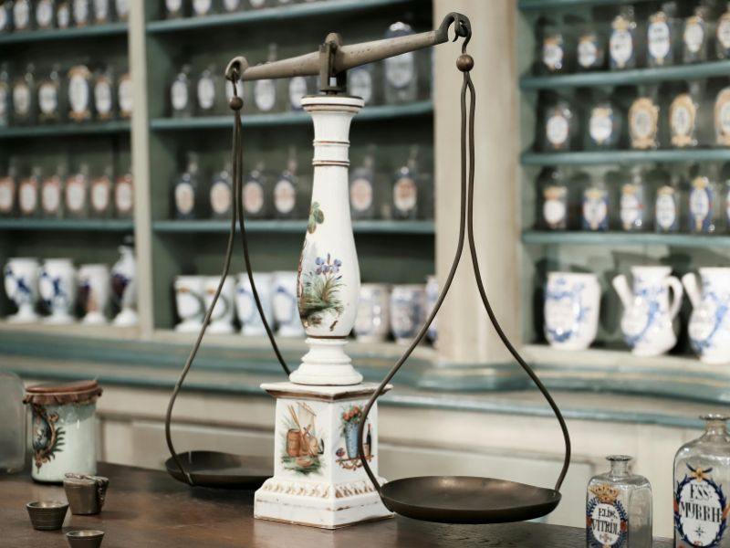 Eine antike Apothekerwaage mit bemalter Keramik und Metallschalen steht auf einem Holztisch vor historischen Apothekerschränken. Dieses Motiv symbolisiert die individuelle Rezepturherstellung und das Streben nach Balance in der Traditionellen Europäischen Medizin.