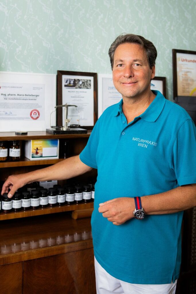 Mag. pharm. Marco Beitelberger steht lächelnd in seiner Praxis neben einem Holzregal mit pflanzlichen Präparaten. Er unterstützt ehrgeizige Klienten mit strukturierten Konzepten dabei, ihre Erschöpfung zu behandeln in Wien.