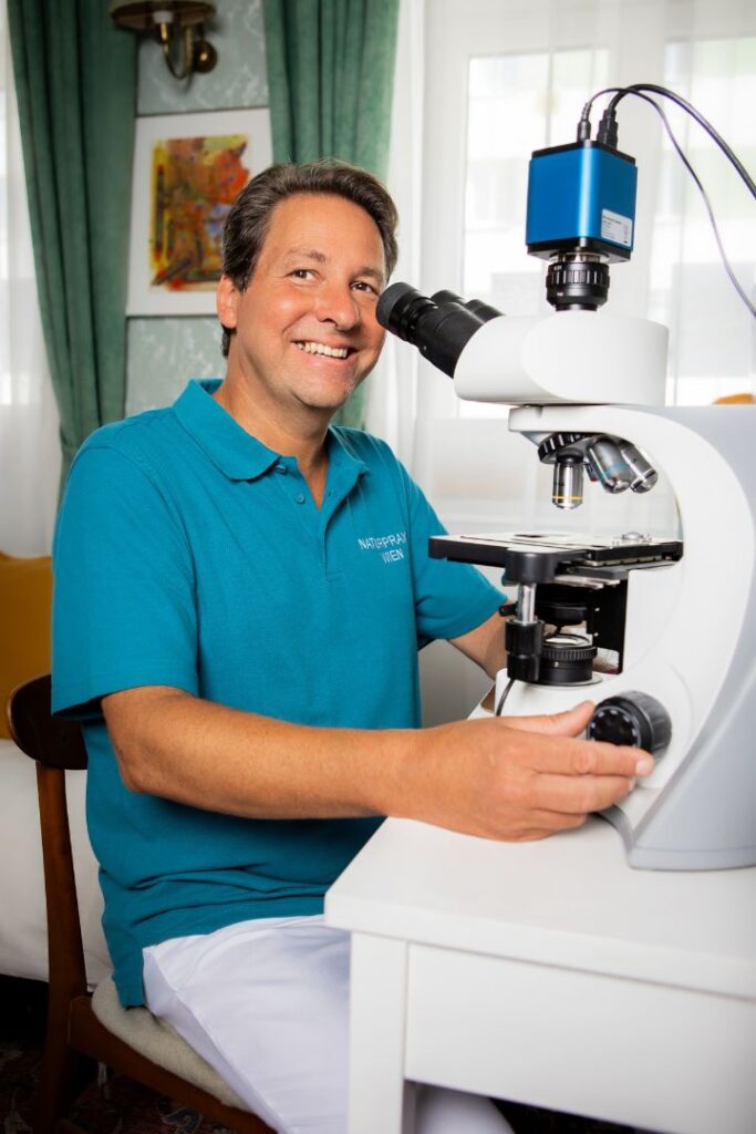 Mag. pharm. Marco Beitelberger sitzt lächelnd an einem modernen Dunkelfeldmikroskop. In seiner Praxis für Naturheilkunde in Wien analysiert er das vitale Blutbild, um den biologischen Ursachen von Erschöpfung und Unwohlsein auf den Grund zu gehen.