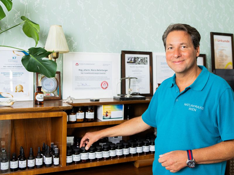 Mag. pharm. Marco Beitelberger steht lächelnd in seiner Praxis für Naturheilkunde in Wien. Er greift nach kleinen Fläschchen mit pflanzlichen Essenzen, die auf einem Holzregal stehen, während im Hintergrund seine fachlichen Zertifikate an der Wand hängen.